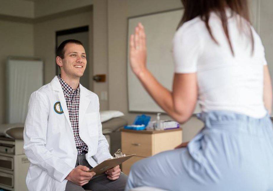 Person in lab coat with clipboard speaking to someone in a medical exam room